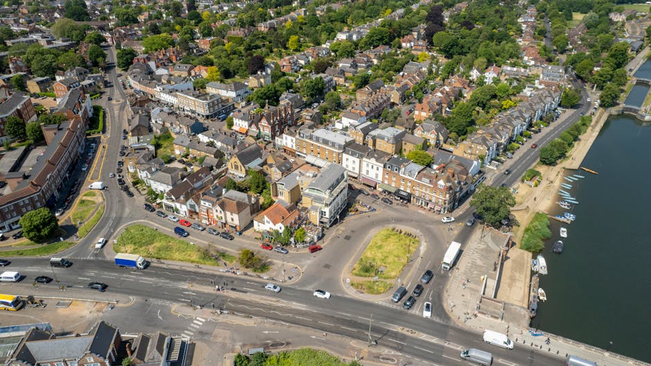Aerial view of a residential area near a body of water, featuring a variety of buildings including shops, houses, and apartment blocks arranged along streets with parked vehicles. The image shows a sm