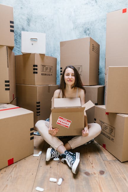 Two women stand indoors amongst a collection of cardboard moving boxes and packing materials, with one holding a medium-sized box labeled with moving stickers and the other holding a decorative wicker